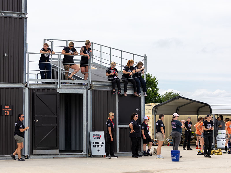Participants gather around a two-story training structure, with several people watching from an upper platform at an outdoor fire service training site.