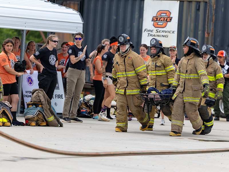 Firefighters in full gear carry a person on a stretcher during a training exercise while others observe at an outdoor fire service training facility.