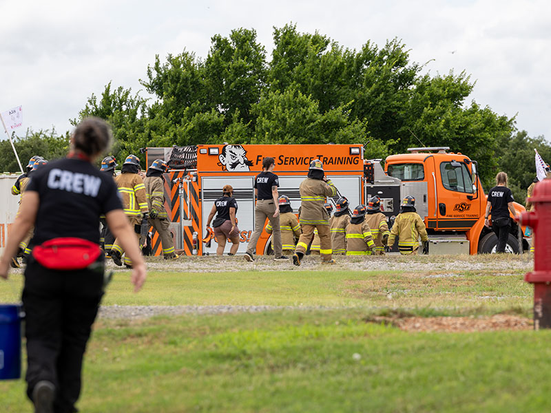 Firefighters and training crew gather around a fire service vehicle during an outdoor training exercise at a fire service facility.