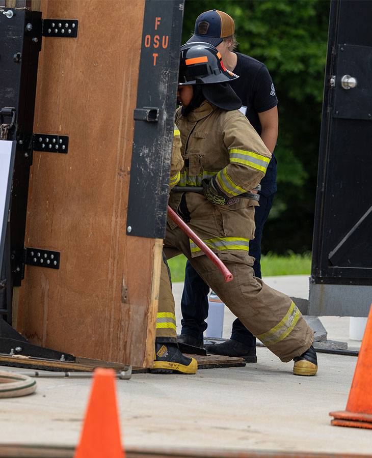 Firefighter in full protective gear uses a pry bar to force open a door during a training exercise at a fire service facility.