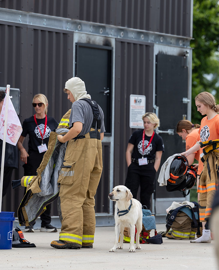 Participants at a fire training site put on turnout gear near a building while a dog stands nearby among equipment.