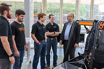 Group of students in matching Cowboy Racing shirts stand with an instructor in a campus lab, discussing a small open‑frame vehicle prototype displayed in front of them.