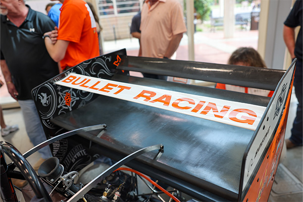 Close-up of the rear wing of a student-built race vehicle labeled “Bullet Racing,” surrounded by onlookers, highlighting the team’s engineering project on display.