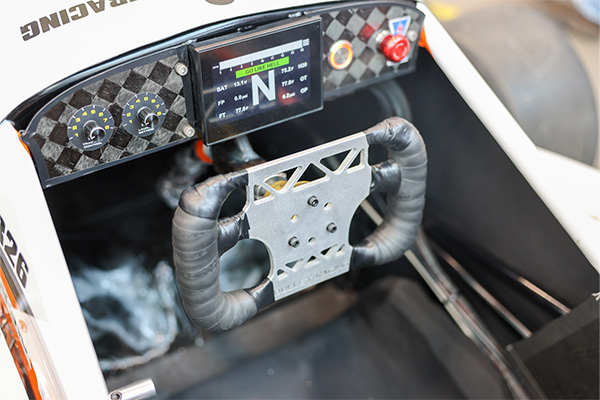 Close-up of a student-built race car cockpit showing a steering wheel and digital dashboard display with gauges and controls.