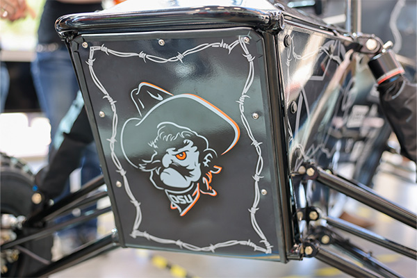 Close-up detail of a small open‑frame vehicle showing a panel with the OSU Pistol Pete logo framed by barbed wire graphics.