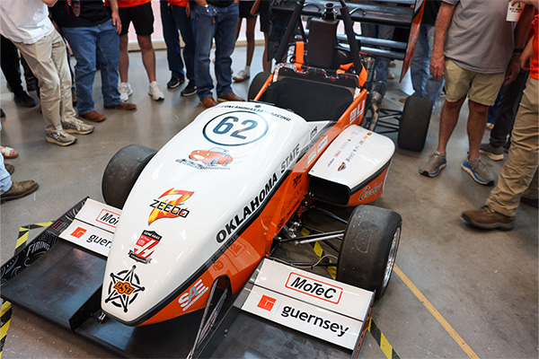 Top view of an Oklahoma State University formula-style race car labeled “62” surrounded by people, highlighting a student-built engineering project on display.