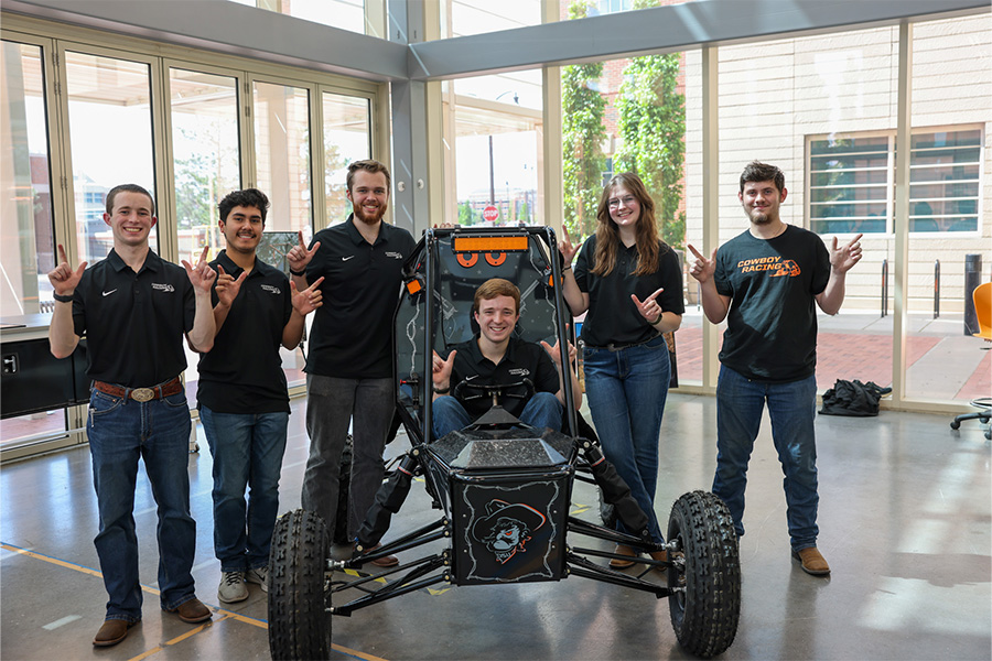 Group of students pose around a student-built off-road vehicle in a campus lab, highlighting a collaborative Cowboy Racing engineering team project.