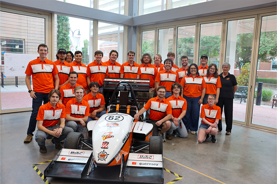 Group of students in matching orange shirts pose around an Oklahoma State University formula-style race car labeled “62” in a campus lab, highlighting a collaborative engineering team and their vehicle.