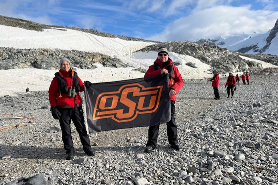Two people in red jackets stand on a rocky, snow-covered landscape holding an Oklahoma State University flag while others in similar gear walk across the glacier in the background.