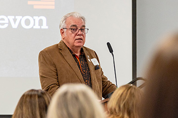 A person stands at a podium speaking into a microphone during a presentation, with a projection screen displaying sponsor text behind them and an audience visible in the foreground.