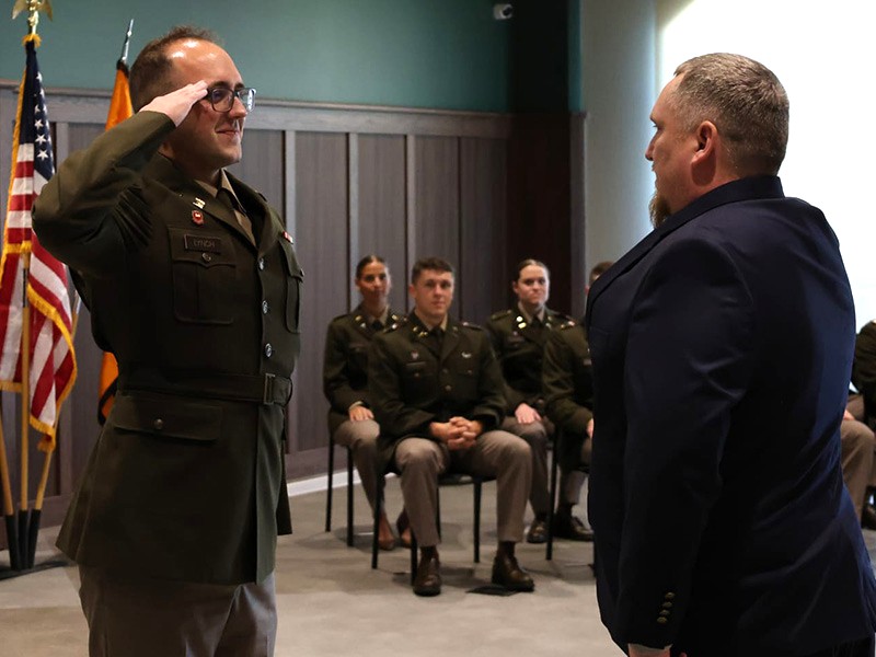 A person in a military uniform raises a hand in salute while facing another individual during a formal ceremony, with seated uniformed attendees and flags visible in the background.