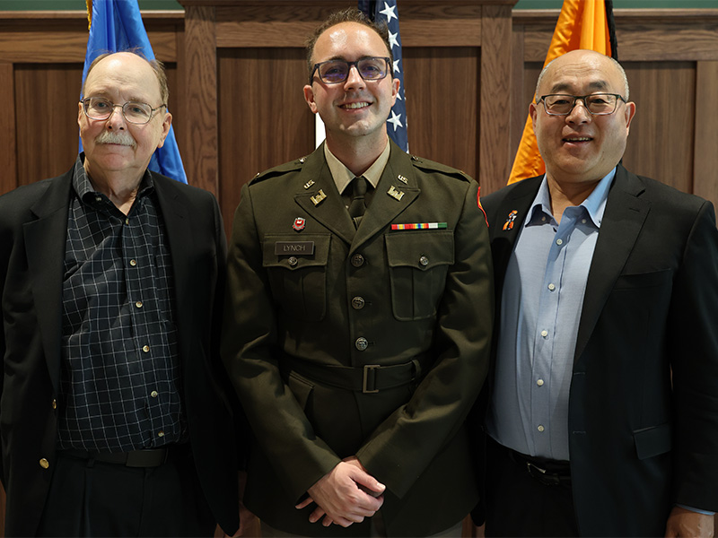 Three individuals, including one in a military uniform, stand side by side in front of flags and a wood-paneled wall, posing for a formal portrait.