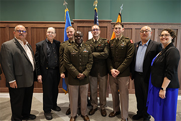A group of individuals, including several in military uniforms and others in business attire, stand together in a formal indoor setting with flags and wood-paneled walls, posing for a group portrait.
