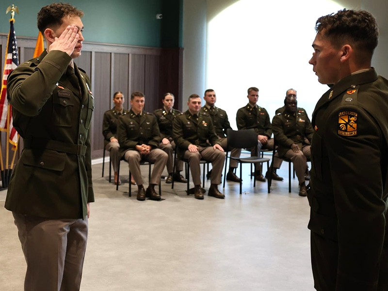 A person in a military uniform raises a hand in salute while facing another uniformed individual during a formal indoor ceremony, with seated attendees and flags visible in the background.