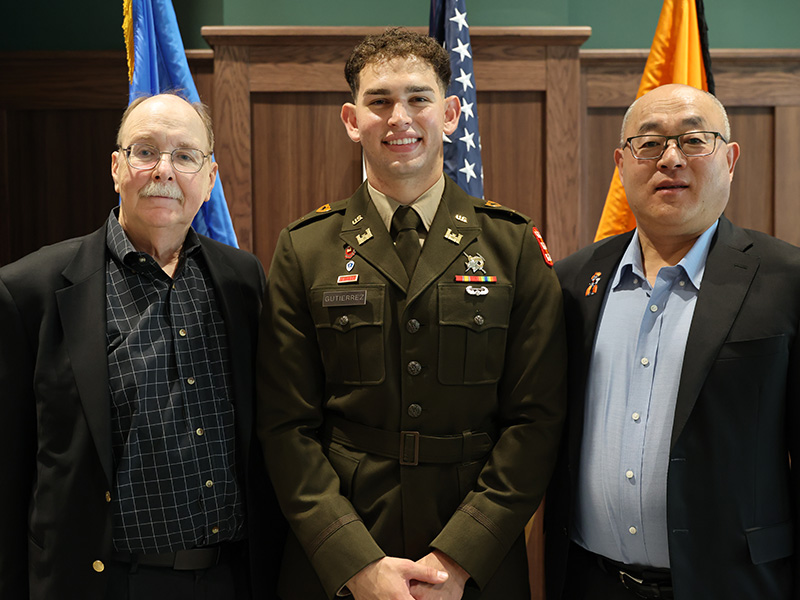 Three individuals, including one in a military uniform, stand in front of flags and a wood-paneled wall, posing for a formal portrait.