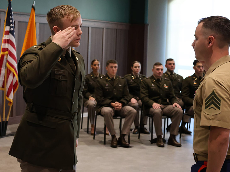A person in a military uniform raises a hand in salute while facing another uniformed individual during a formal indoor ceremony, with seated attendees and flags visible in the background.