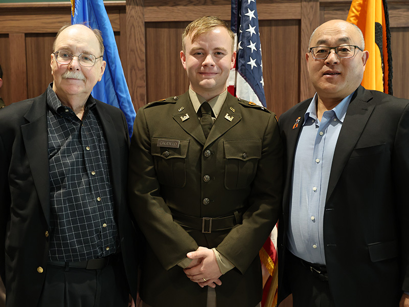 Three individuals, including one in a military uniform, stand side by side in front of flags and a wood-paneled wall, posing for a formal portrait.