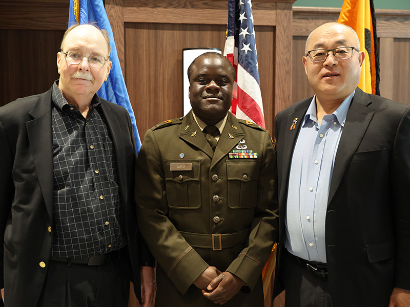 Three individuals, including one in a military uniform, stand side by side in front of flags and a wood-paneled wall, posing for a formal portrait.