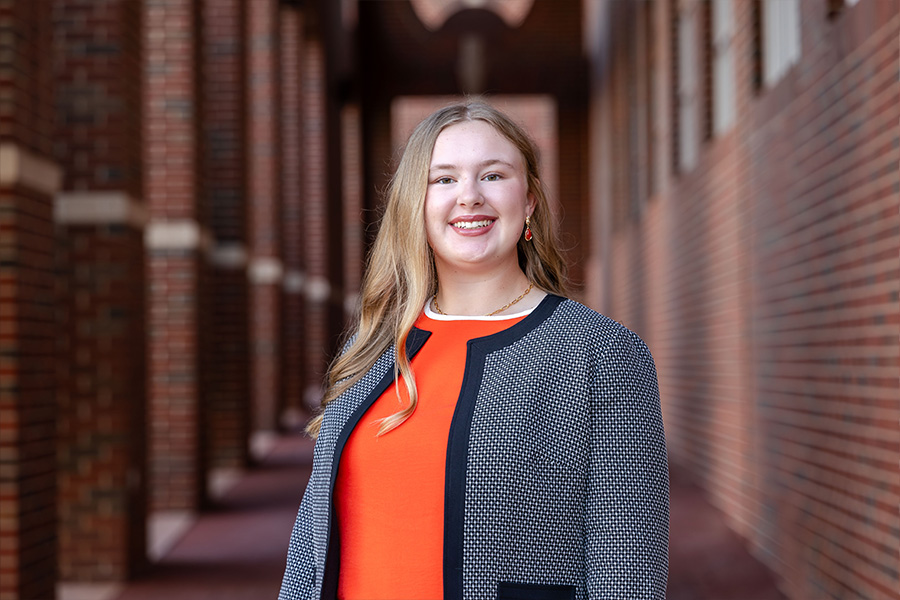 Girl with long blond hair wearing a gray blazer and orange shirt stands in front of a brick building