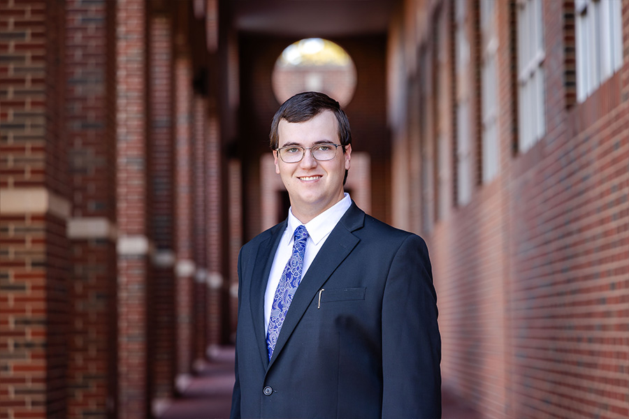 Man with a black suit and blue tie stands in front of a brick building