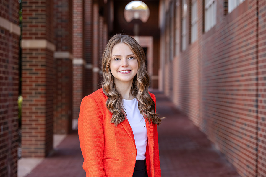 Girl with long hair wearing an orang blazer and white dress shirt stands in front of a brick building