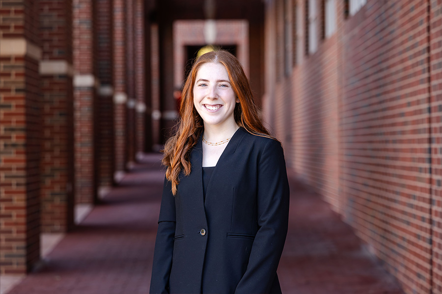 Girl with long hair wearing a black blazer stands in front of a brick building