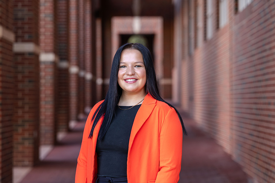Girl with long hair wearing an orange blazer and black shirt stands in front of a brick building