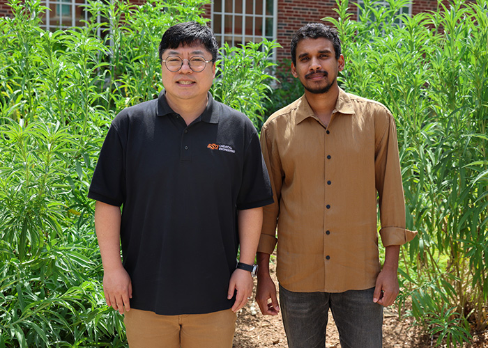 Two men stand side by side outdoors in front of tall, leafy green plants.