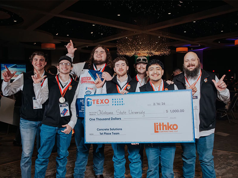 A group of college students wearing medals and name badges celebrate on stage while holding a large first‑place competition check, highlighting their team’s win at a concrete industry student competition.