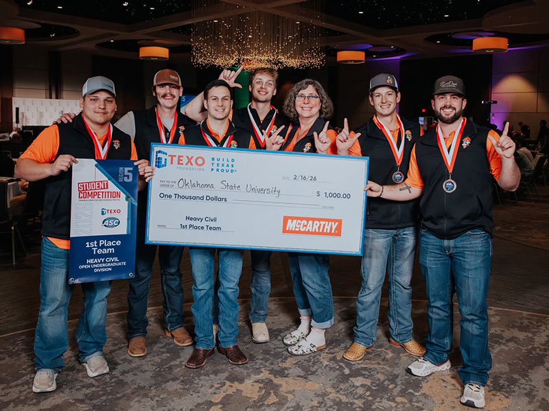 A group of students wearing medals stand together holding a large first‑place competition check, celebrating Oklahoma State University’s Heavy Civil team victory at a student construction competition.