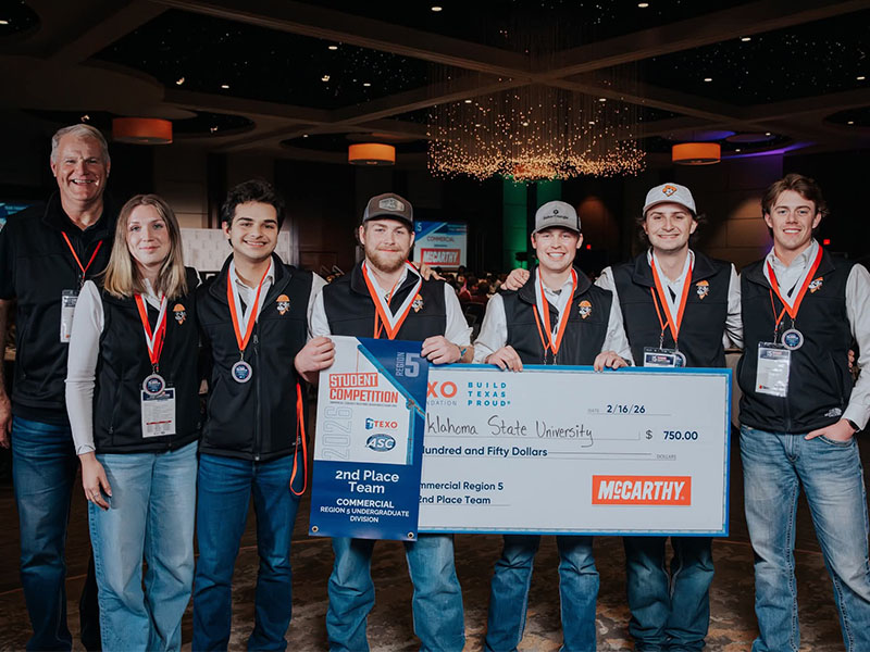 A group of students wearing medals pose together while holding a check and banner recognizing a second‑place finish at a commercial construction student competition.