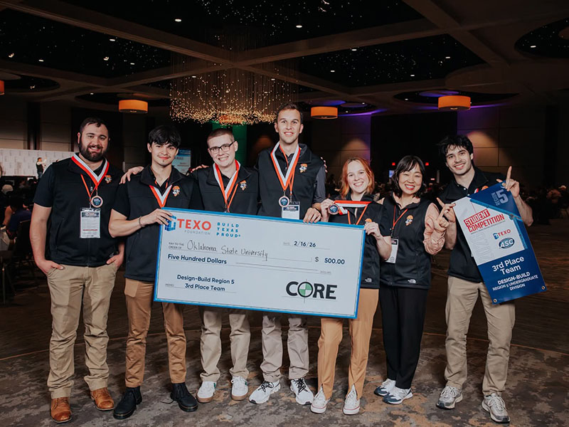 A team of college students wearing medals pose together while holding a check and a third‑place banner, recognizing their achievement in a design‑build category at a student construction competition.