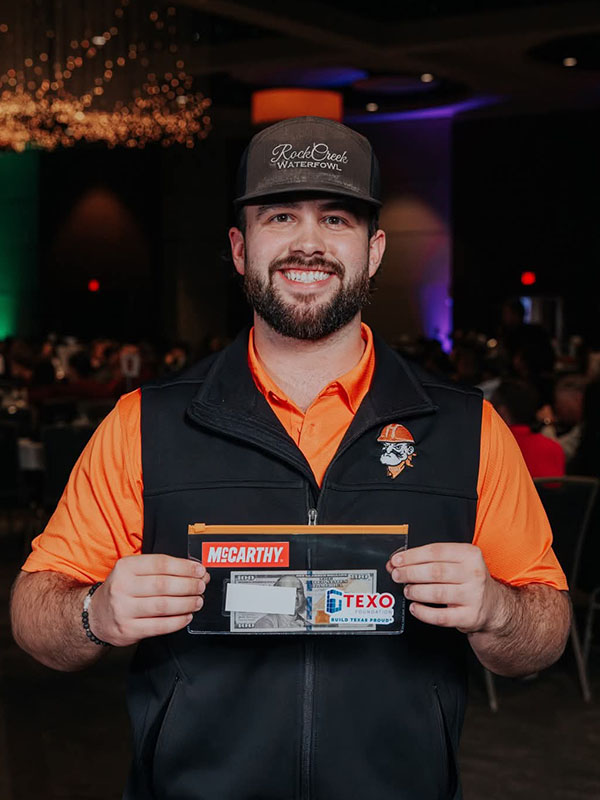 A smiling student wearing a cap and Oklahoma State University vest holds an award envelope at an industry‑sponsored student competition, highlighting individual achievement and recognition.