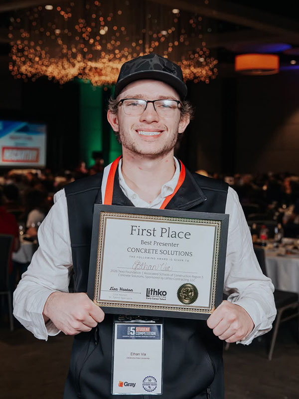 A smiling student wearing a name badge holds a framed certificate for first place best presenter at a student competition, highlighting academic achievement and recognition at a professional event.