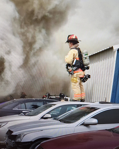 A firefighter wearing full turnout gear and a self-contained breathing apparatus stands outside a smoke-filled building during an active fire response
