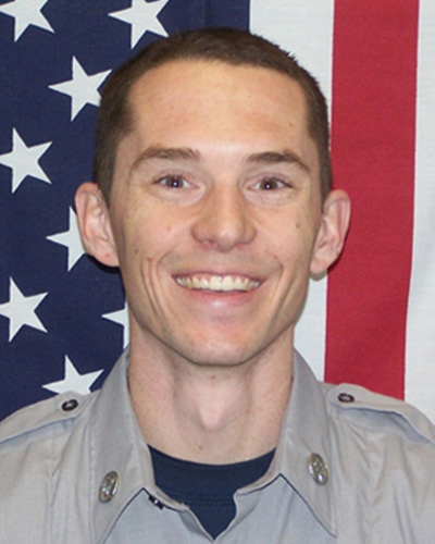 head-and-shoulders portrait of a smiling man wearing a light gray emergency services uniform in front of an American flag backdrop