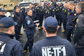 A group of approximately 30 members of Nebraska Task Force 1 Urban Search and Rescue stand in a parking lot during a briefing before deployment.