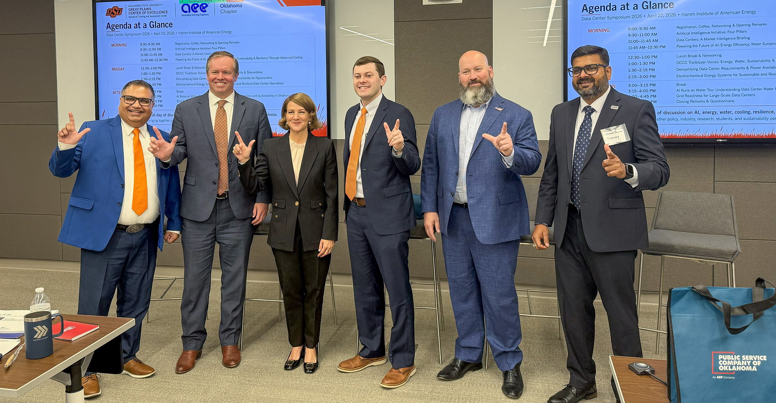 Color photograph of six people in business attire standing indoors in front of presentation screens displaying a conference agenda, posing together and making hand gestures.