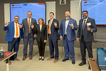 Color photograph of six people in business attire standing indoors in front of presentation screens displaying a conference agenda, posing together and making hand gestures.