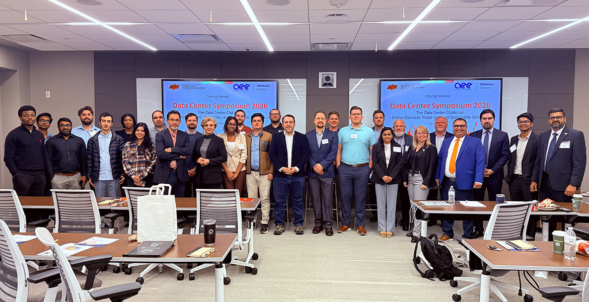 Wide-angle color photograph of a group of conference attendees standing together in a meeting room, with presentation screens behind them displaying “Data Center Symposium 2026” and tables with materials in the foreground.