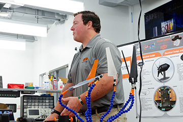 The image features a male individual standing in a workshop or laboratory environment, engaged in presenting or teaching