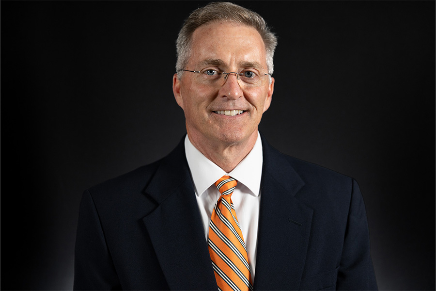 Man with glasses in a black suit with orange tie smiles for portrait