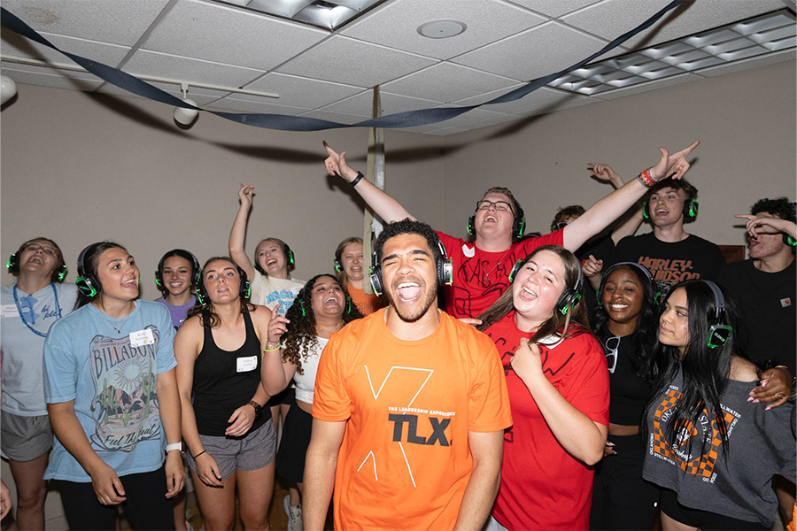 A large group of participants wearing over‑ear headphones stand closely together in an indoor room, some raising their arms and moving as if dancing during a silent‑disco style group activity.