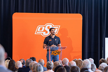 A speaker stands at an Oklahoma State University podium addressing an audience, with an orange OSU backdrop behind and a presentation screen to the right displaying the name “Jacob Smith” and leadership titles.