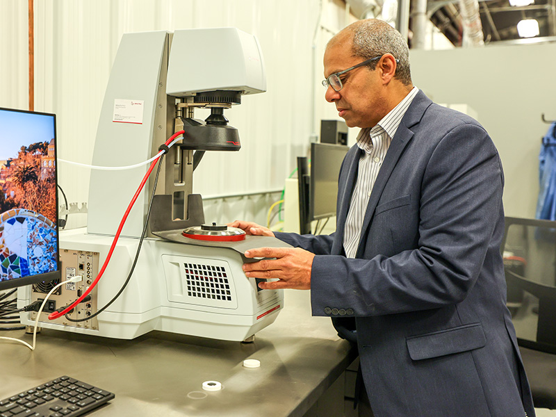 Researcher operating a materials testing instrument in a laboratory, with a computer monitor and analytical equipment visible on the workbench.