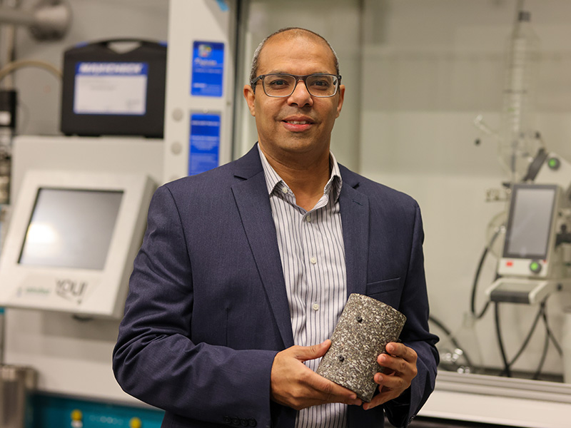 Researcher holding a cylindrical material sample in front of laboratory testing equipment.