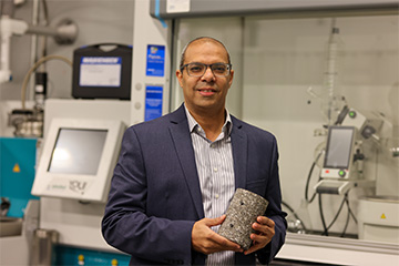 Researcher holding a cylindrical material sample in front of laboratory testing equipment.