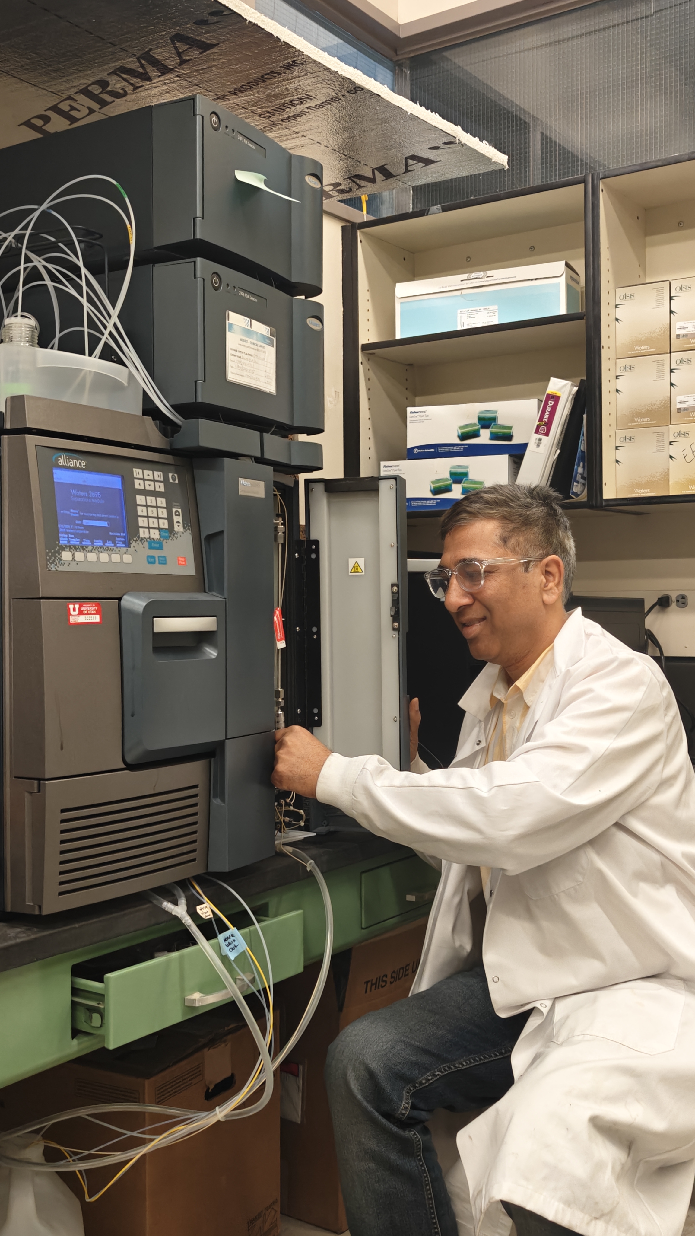 A researcher in a laboratory adjusts equipment inside an analytical instrument, with tubes and cables connected to the machine and shelves of lab supplies in the background.