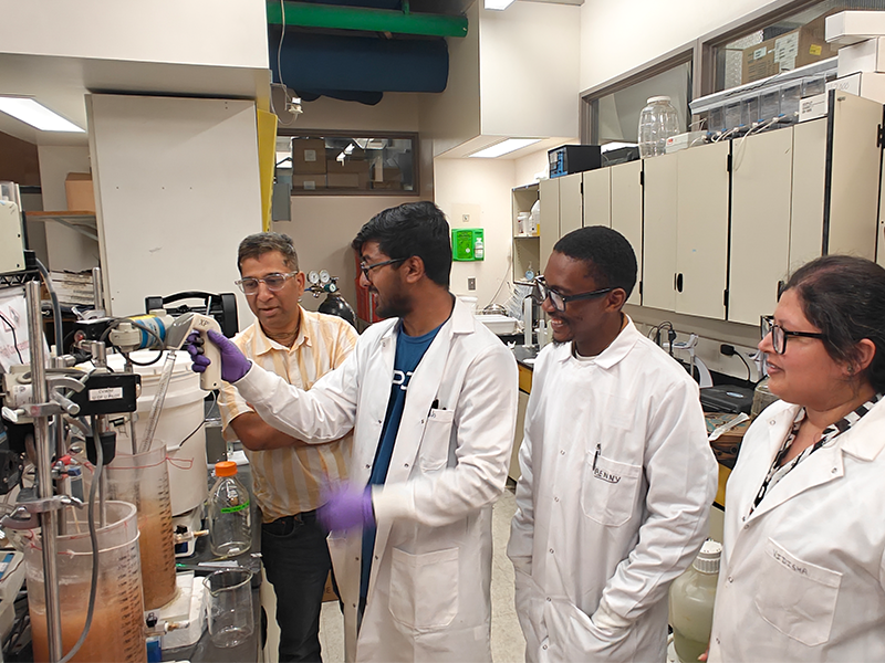 Four researchers in lab coats gather around laboratory equipment as one researcher operates a probe and tubing system connected to containers of liquid while the others observe in a research laboratory.