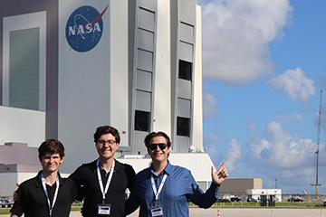 The image captures three young men standing in the foreground, smiling and posing for the camera in front of a large NASA facility.